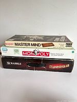 Stacked view of three boxed board games: Super Master Mind, Monopoly, Scrabble Deluxe