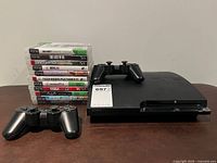 Front view of PS3 Slim console with two controllers and stacked game cases