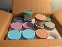 Open carton showing multiple pastel-lidded glass jars seated in foam