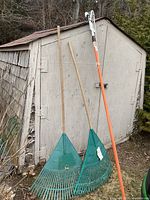 Two green fan rakes and orange pole pruner leaning against shed
