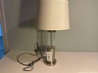 Full view of lamp showing glass column, chrome base, white shade, power cord