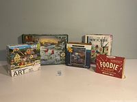 All four jigsaw puzzle boxes and Foodie Fight game box displayed upright on table