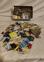 pile of assorted matchbooks spread on surface with tin in background
