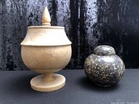 Tan pedestal urn and dark speckled rounded urn side by side, lids on