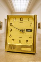 Front view of Citizen quartz desk clock showing gold-tone case, yellow dial, black numerals, ornate hands, day-date window, speaker grille