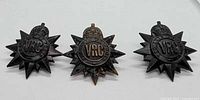 Front view of three Victoria Rifles of Canada cap badges showing starburst design, crown, and VRC roundel