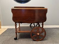 Side view of mahogany drop-leaf tea cart showing carved skirt, large spoked wheel, caster wheels, lower shelf