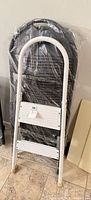 Folded white 2-step ladder leaning against plastic-wrapped black folding chair