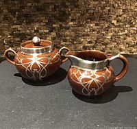 Front view of brown Denby sugar bowl with lid and matching creamer showing silver overlay pattern