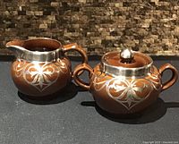 Front view of brown glazed Denby creamer and sugar bowl showing silver overlay design