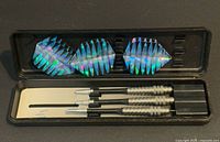 Open black case showing three silver-barrel darts and three loose blue holographic flights