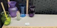 Five glass vases and bottles lined on table with rulers for scale