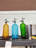 Three coloured seltzer bottles and metal shaker on shelf