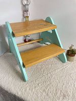 Front angled view showing both steps, side panel shape, turquoise paint, dowel stretcher, small planter for scale