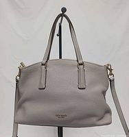 Front view of taupe Kate Spade Abbott small satchel on stand showing pebbled leather, dual handles, logo, crossbody strap attached