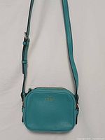 Front view of turquoise leather camera crossbody bag showing logo and strap
