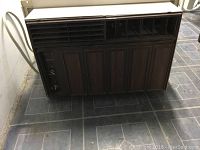 Full view of the older, dark brown air conditioner unit on tiled floor showing vent grill and wood-paneled lower front.
