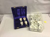 Wide view of the egg cups lot showing four decorated egg cups in a purple velvet case beside a plastic container holding multiple plain white egg cups.