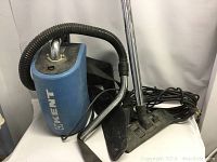 Kent brand blue commercial backpack vacuum with hose, metal wand, and floor nozzle on white surface with visible wear and scuffs.