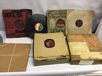 Approximately 25 vintage 78 RPM records with various paper sleeves including branded Rex and Parlophone sleeves, stacked vertically and spread across a surface.