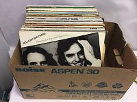 Approximately 50 mixed genre LP records stored in a recycled copy paper box. The front visible record is by Bellamy Brothers featuring 'Let Your Love Flow'. Other album covers partially visible behind.