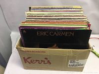 Box holding approximately 50 vinyl LP records, showing Eric Carmen album at front with other records behind in mixed condition.
