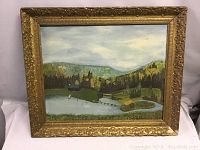 Full view of signed oil painting depicting a lake cottage scene with surrounding greenery in a gold ornate frame.