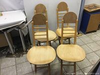 Four folding wooden chairs arranged together, showing the front and side profiles with visible backrests and seats.