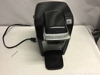 Full view of black Keurig coffee maker with silver front panel and cup holder section. Power cord visible.