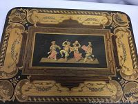 Top view of the music box lid showing detailed inlay with figures and ornamental designs in wood and black background, decorative border.