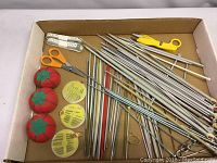 Box with assorted knitting needles, crochet hooks, orange scissors, yellow thread cutter, tomato pin cushions, and needle cases with sewing needles