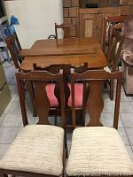 Photo showing the solid oak table with the leaf extension and the backs of six chairs around it. Chair seat colors/textures vary.