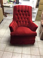 Front view of red velvet upholstered rocker chair showing tufted backrest, angled armrests with pleated fabric, cushioned seat, and fabric skirt base.