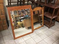 Two wooden framed wall mirrors standing on floor against shelving, large and smaller mirror visible with plain and decorative top frames respectively.