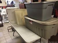 Four plastic containers including two large storage containers stacked on the white table and two plastic laundry baskets behind them.