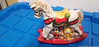 Front view of white ceramic rocking horse piggy bank with toys at base