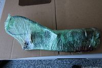 Grease gun fully wrapped in green plastic film showing overall shape and attached hose