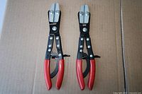 Two identical red-handled hose clamp pliers laid flat on cardboard