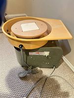 Front view of potter's wheel showing metal housing, yellow splash pan, wheel head, foot pedal, power cord