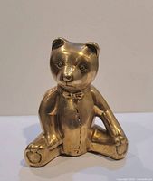 Front view of brass teddy bear coin bank showing seated pose, bowtie, stitched belly