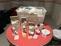 Large plastic tote filled with boxed Precious Moments figurines, three unboxed figurines, mug, plate and plush doll set on table