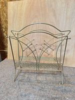 Front view of metal magazine stand showing crossed rod sides, arched handle, lower shelf.