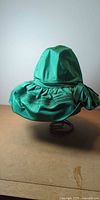 Front view of emerald satin hat on stand showing crown and ruched brim