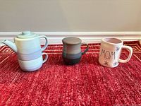 Three ceramic drinkware pieces lined up: teapot with cup, gray-green mug, MOM mug