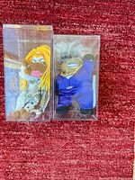 Both plush figures side by side in clear cases on red surface