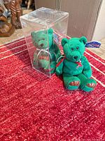 Loose green holiday plush teddy bear and second identical bear inside clear plastic display box on red rug