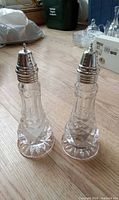 Full view of both clear pressed glass shakers standing upright on wood floor