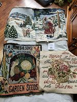 All four tapestry pillow fronts laid out on floor: two winter scenes, garden seeds design, floral basket script