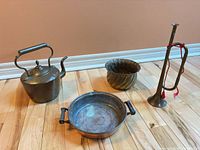 Group shot of brass kettle, brass bugle with red cord, copper pan with handles, and ribbed brass planter on hardwood floor