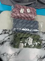 Three sealed plastic packs of cheesecloth table runners in dusty rose, slate blue, sage green